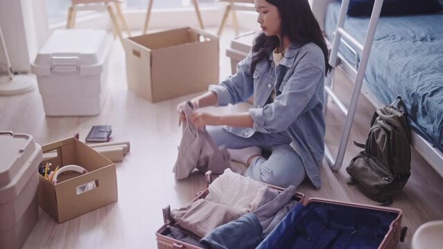 young Asian student in casual clothing is seen unpacking her luggage, taking out clothes and neatly organizing them in her dorm room. She efficiently arranges her belongings in the living space.