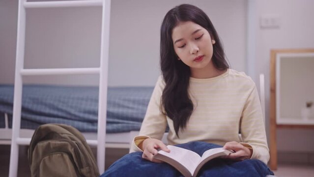 young Asian student in casual clothing sits on her bunk bed in a dorm room, engrossed in a book. She rests against the raised backrest of her bunk bed in her cozy dormitory.