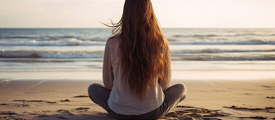 Young woman with long hair sitting on the beach seen from behind in an athletic pose with her hands resting on her knees - obrazy, fototapety, plakaty
