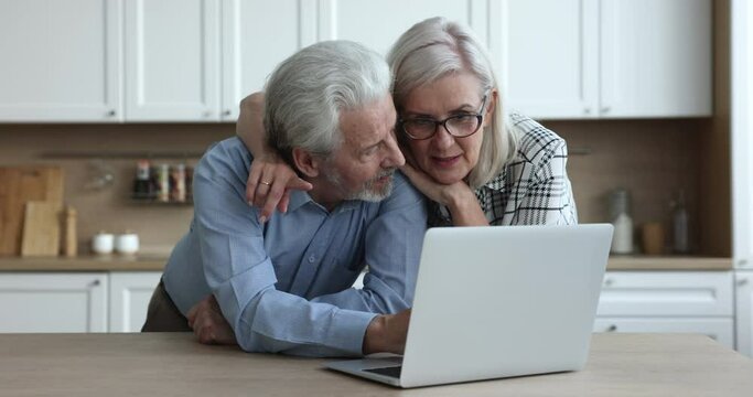 Happy senior married couple enjoying modern Internet communication at home, shopping together at laptop, making online order, using healthcare medical service on Internet