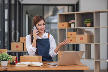 A portrait of a young Asian woman, e-commerce employee sitting in the office full of packages in the background write note of orders and use smartphone, for SME business ecommerce and delivery