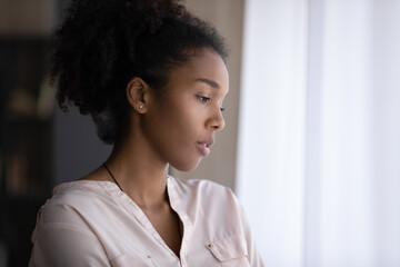 Thoughtful sad millennial Black d girl looking out of window in deep thoughts, thinking over...