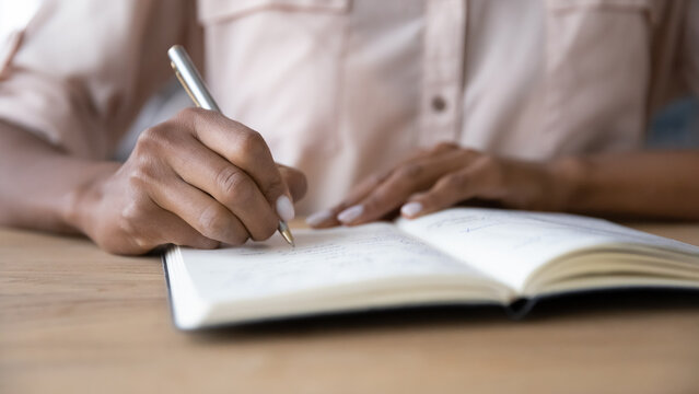 Hands of dark skinned student girl, writer, author making notes in notebook with pen, writing summary of webinar, draft of essay. Woman planning work tasks, using organizer, workbook. Close up