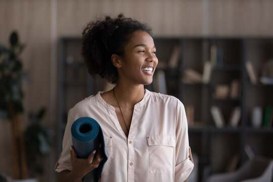 Happy Black teen girl holding rolled yoga mat, preparing for meditation, morning workout, exercise at home. Young millennial woman enjoying active lifestyle, staying fit, looking away, smiling