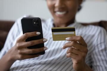 Hands of happy black young shopping woman holding credit card and smartphone, using banking app, virtual service, mobile phone for payment, paying for purchase, order, booking on internet. Close up - obrazy, fototapety, plakaty
