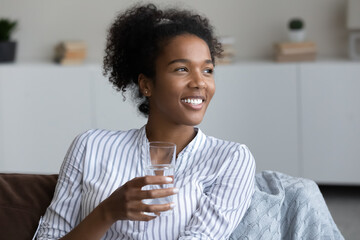 Happy young African woman, millennial girl holding glass of pure fresh clean water, drinking,...