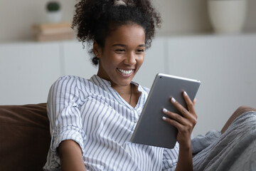 Happy excited millennial girl, young Black woman reading book on tablet computer, watching funny movie, making video call, talking at pad webcam, relaxing on sofa at home, smiling, laughing - obrazy, fototapety, plakaty