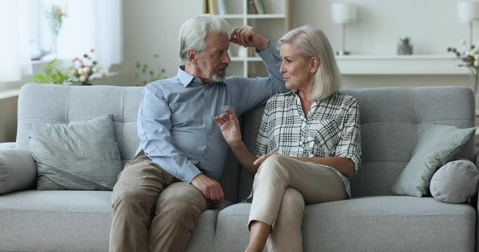 Serious elderly dating couple sitting on sofa at home, talking, getting positive. Blonde senior wife talking to grey haired husband on couch, discussing retirement, enjoying family leisure