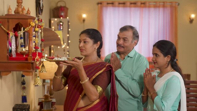 Denotational Mother offering aarthi to god with praying father and daughter during diwali pooja rituals at home - Concept of religious custom, traditional bonding and family rituals