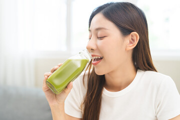 Green detox juice concept, Beautiful smile asian young woman hand holding vegetable smoothie bottle for diet at home. Happy girl drinking healthy meal food for weight loss. Lifestyle, vegan nutrition. - obrazy, fototapety, plakaty