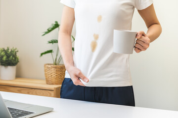 Cloth stain, disappointment asian young woman, girl clumsy with hot coffee, tea stains, hand show making spill drop on white t-shirt, spot dirty or smudge on clothes while working on computer at home. - obrazy, fototapety, plakaty