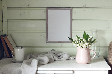 spring mockup with an empty frame on the wall, a white mug and a bouquet of lilies of the valley.