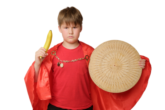 Funny child boy in a medieval knight costume, isolated on background. Boy in a red shirt and a straw shield holds a banana in his hands