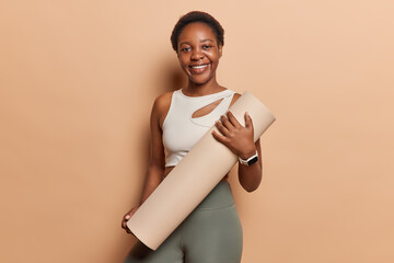 Smiling active woman dressed insportsclothes carries rolled karemat going to practice yoga or...