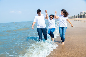 Happy indian Family of three enjoying summer vacation. Man and woman holding hands of their daughter and walking on beach. - obrazy, fototapety, plakaty