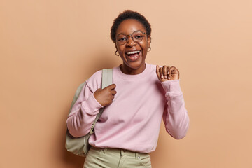 People positive emotions concept. Indoor waist up of young happy smiling broadly African american female student standing in centre on beige background wearing casual clothes and bag on shoulder - obrazy, fototapety, plakaty