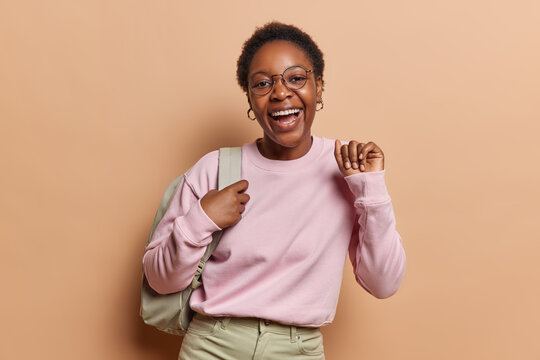 People positive emotions concept. Indoor waist up of young happy smiling broadly African american female student standing in centre on beige background wearing casual clothes and bag on shoulder