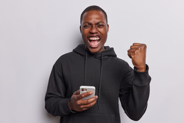 Emotional dark skinned guy feels like winner clenches fist and exclaims loudly trading or shopping while sale season reacts to something wears casual black sweatshirt isolated over white background - obrazy, fototapety, plakaty