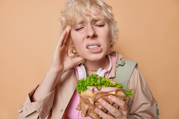 Cheat meal day concept. Upset woman suffers from headache keeps hand on temple clenches teeth eats...
