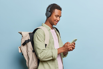 Sideways shot of curly haired African male student listens music via stereo headphones holds mobile phone carries big rucksack on back poses against blue background. Modern lifestyle concept