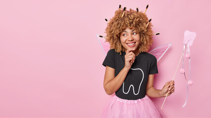 Positive curly haired woman pretends being tooth fairy poses with magic wand dressed in costume comes on carnival focused aside happily isolated over pink background copy space for your promotion