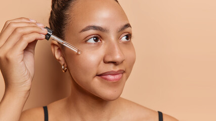 Beauty procedure. Studio close up of young beautiful cheerful smiling gently European female standing isolated on beige background in black undershirt applying acid oil for face looking aside