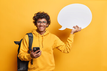 Indoor shot of curly haired Hindu man carries rucksack on shoulder demonstrates blank speech bubble for your advertising content uses smartphone for online communication wears spectacles sweatshirt - obrazy, fototapety, plakaty