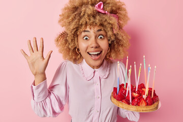 Holiday celebration concept. Studio photo of young beautiful cheerful smiling broadly African american woman standing in centre isolated on pink background holding cake with strawberries and candles - Powered by Adobe