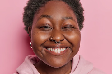 Close up shot of African woman with short curly hair bites lips smiles toothily shows white teeth winks eye expresses positive emotions has playful expression poses against pink studio background