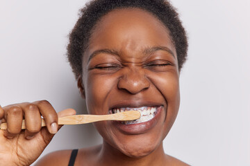 Teeth hygiene concept. Close up shot of dark skinned woman with curly hair cleans teeth holds wooden toothbrush has eyes closed isolated over white background. Morning procedure and perfect smile - obrazy, fototapety, plakaty