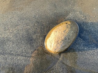 A rock rounded with wet sand in river water, beach in sun light