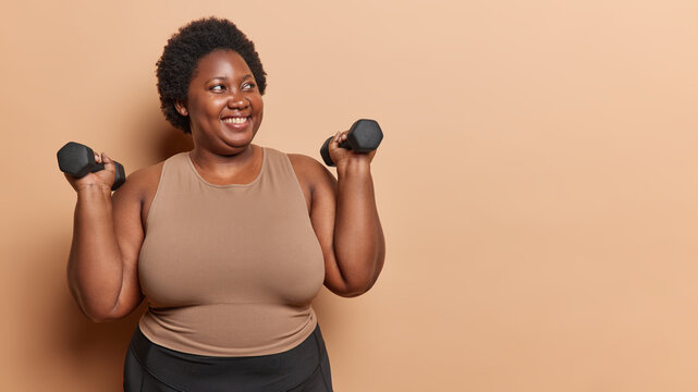 Body positive dark skinned African woman lifts dumbbells has hard training dressed in sportswear goes in for sport in gym looks happily aside poses against brown background copy space for your text
