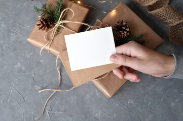 Christmas presents, gift boxes wrapped with brown crafted paper, pine cones and twig decor, on gray table background. Empty paper card mockup in hand. Gift tag, business card or invitation template