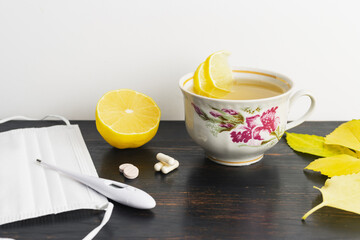 Tea cup with lemon, thermometer, pills and medical mask on a wooden table with yellow leaves. Concept of treatment autumn cold, flu and coronavirus