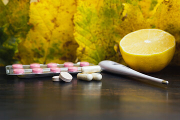 Capsules, pills, medicine in a blister pack, thermometer, lemon on a wooden table with yellow leaves. Concept of treatment autumn cold, flu and coronavirus