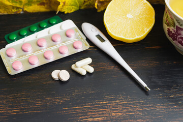 Capsules, pills, medicine in a blister pack, thermometer, lemon on a wooden table with yellow leaves. Concept of treatment autumn cold, flu and coronavirus