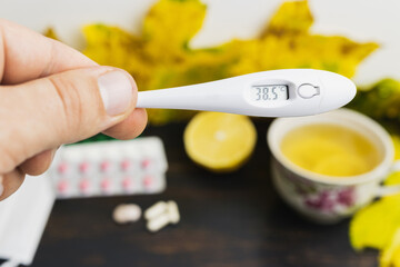 Male hand holds digital thermometer indicates high temperature 38.5, above table with tea cup, lemon, capsules, pills and yellow leaves. Concept of treatment fever, autumn cold, flu and coronavirus - obrazy, fototapety, plakaty
