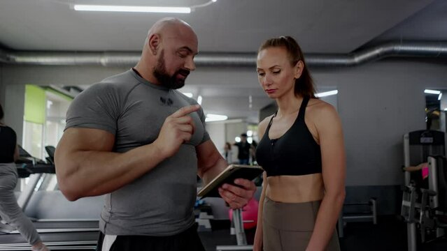 A man fitness trainer and a girl client are talking, smiling and agreeing on schedule for visiting sports facility. The sport coach uses tablet, gives recommendations to the woman athlete to increase