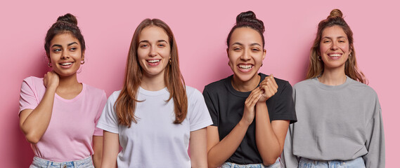 Four cheerful young women express positive emotions and feelings being in good mood smile broadly dressed in casual clothing being best friends isolated over pink background. Set of happy people