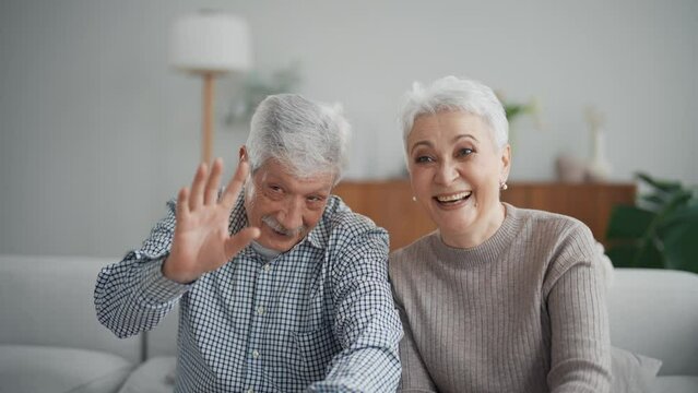 Smiling happy senior couple man and woman calling video chat and emotionally talking looking at camera. They wave hands greeting talker. Modern technologies in everyday life of old people concept.