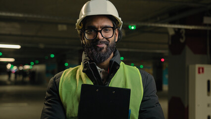 Indian contractor mechanic guy in helmet and glasses smile posing with clipboard folder. Arabian...