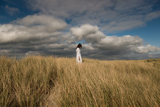 Art portrait of woman in white dress standing alone in field of grass in dunes under cloudy sky