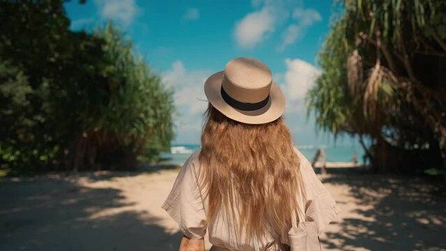 Romantic blonde woman tourist in straw hat walking to ocean sandy beach in sunny day, rear view. Female lady enjoying nature on vacations summer holidays. Travel, tourism, journey, rest relax concept.