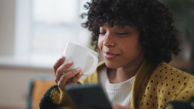Close up young woman with phone drink tea African American woman hold in hand use mobile phone drink coffee Girl relax spend free spare time in living room with phone. Communication on social networks