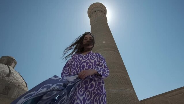 A young woman in a white dress and a long oriental robe dances in the sunshine next to an ancient minaret