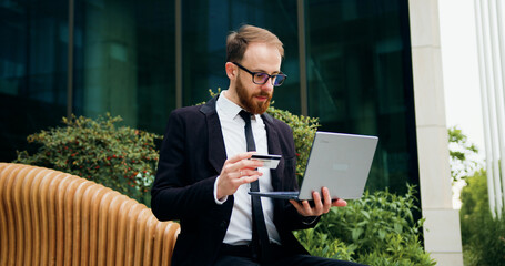 Young businessman using laptop buy on internet by credit card. Cheerful man, laptop and credit card for online shopping, ecommerce or purchase sitting on bench outdoor.