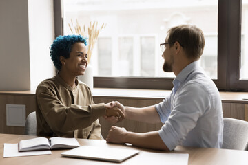 Cheerful couple of office colleagues shaking hands, laughing, celebrating successful teamwork, job result, partnership, agreement. Happy African client giving greeting handshake to manager