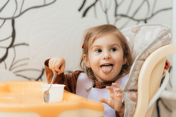 Small cute little toddler brunette caucasian girl with two tails tasting and enjoying by herself with a spoon the greek yogurt sitting in baby chair with messy face self-feeding concept