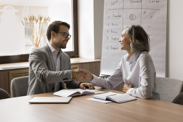 Happy mature businesswoman giving handshake to younger colleagues man, discussing agreement at meeting table. Employer and candidate shaking hands after successful job interview - obrazy, fototapety, plakaty