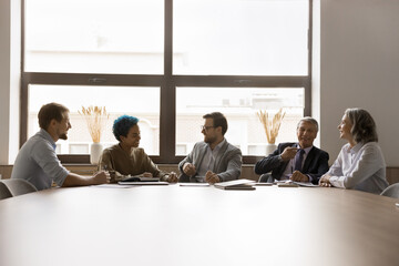 Multiethnic younger and elder business coworkers sitting in row together at large meeting table, talking, laughing, enjoying informal conversation, communication, networking